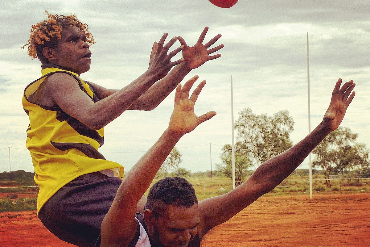Two men, Vernon Japaljarri Dickson and Simon Japanangka Fisher Jr. are both trying to grab a football out of the air while wearing football jerseys. Behind them are football goals set in a red dirt field.